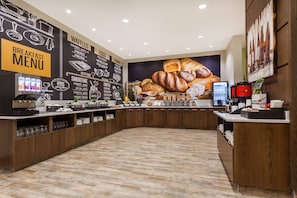Un espace dédié au petit-déjeuner, avec un tableau affichant le menu, des machines à café et une variété d’aliments présentés sur le comptoir.
