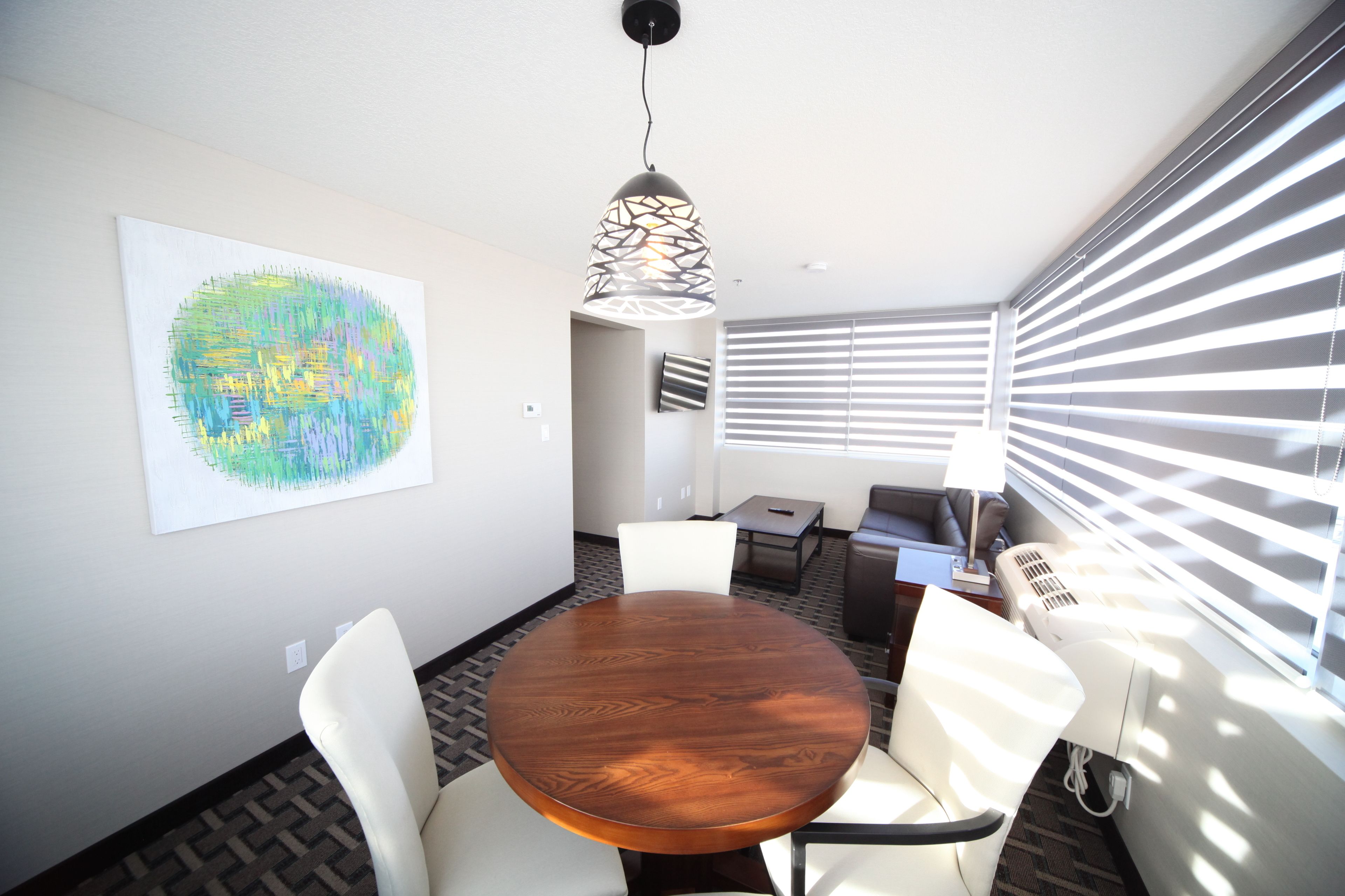 A conference room with a wooden table, white chairs, a hanging pendant light, and a large abstract painting on the wall.