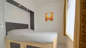 A bedroom with a wooden bed frame, a white mattress, a headboard, a small wall-mounted shelf, and a window with curtains. - Holiday Home Koewacht near Seaside (Koewacht)