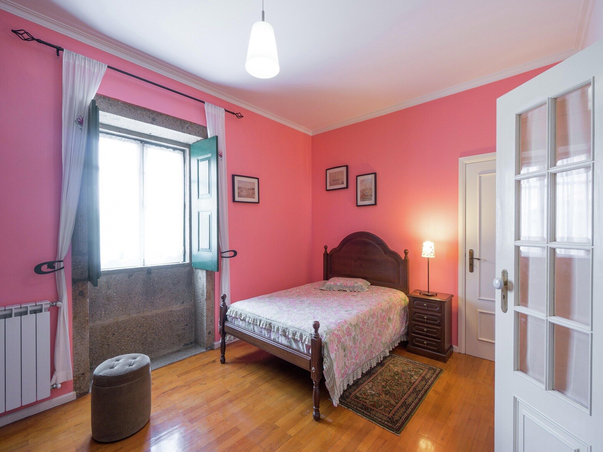 A bedroom with a bed, a nightstand, a lamp, a window with curtains, a radiator, and a door.