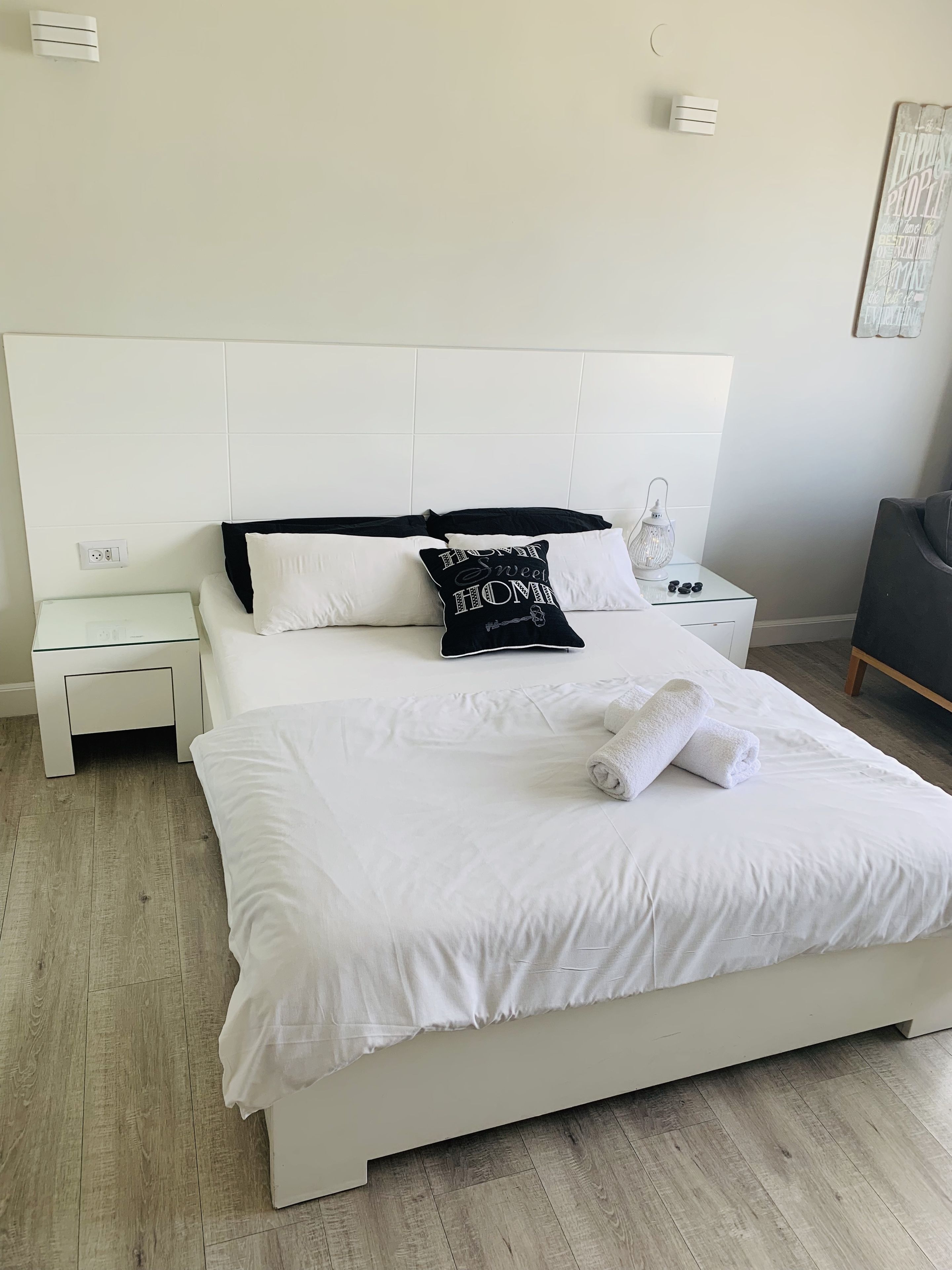 Ein ordentlich bezogenes Bett mit weißer Bettwäsche und einem schwarzen Kopfkissen mit Text, ein Nachttisch mit Lampe und eine Wand-Klimaanlage.
