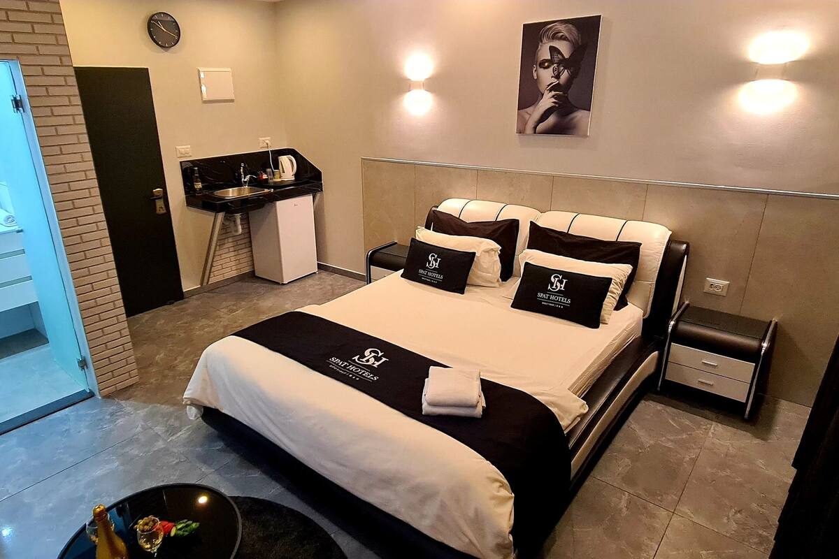 A modern hotel room with a large bed, a small kitchenette, a built-in mirror, and a bathroom with a shower and toilet.