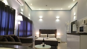 A modern hotel room with a bed, a sofa, a round table, a minibar, and a microwave.
