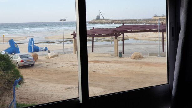 A beach view from inside a building, featuring a sandy beach, a pier, and a car parked near a fence.