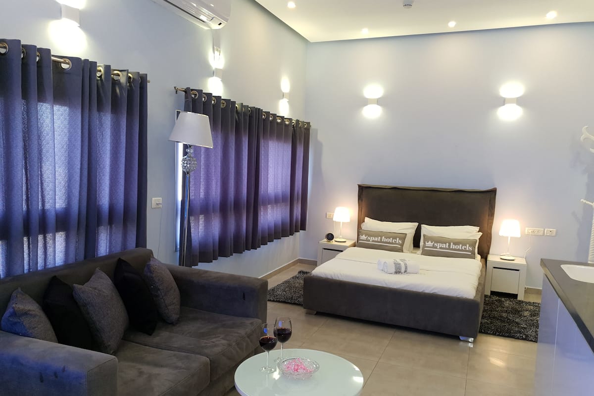 A modern hotel room with a sofa, a bed with branded pillows, a small table with glasses, and a nightstand with a lamp.