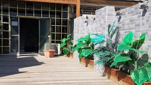 A wooden deck leading to a building with a glass door, surrounded by large green plants in wooden planters.