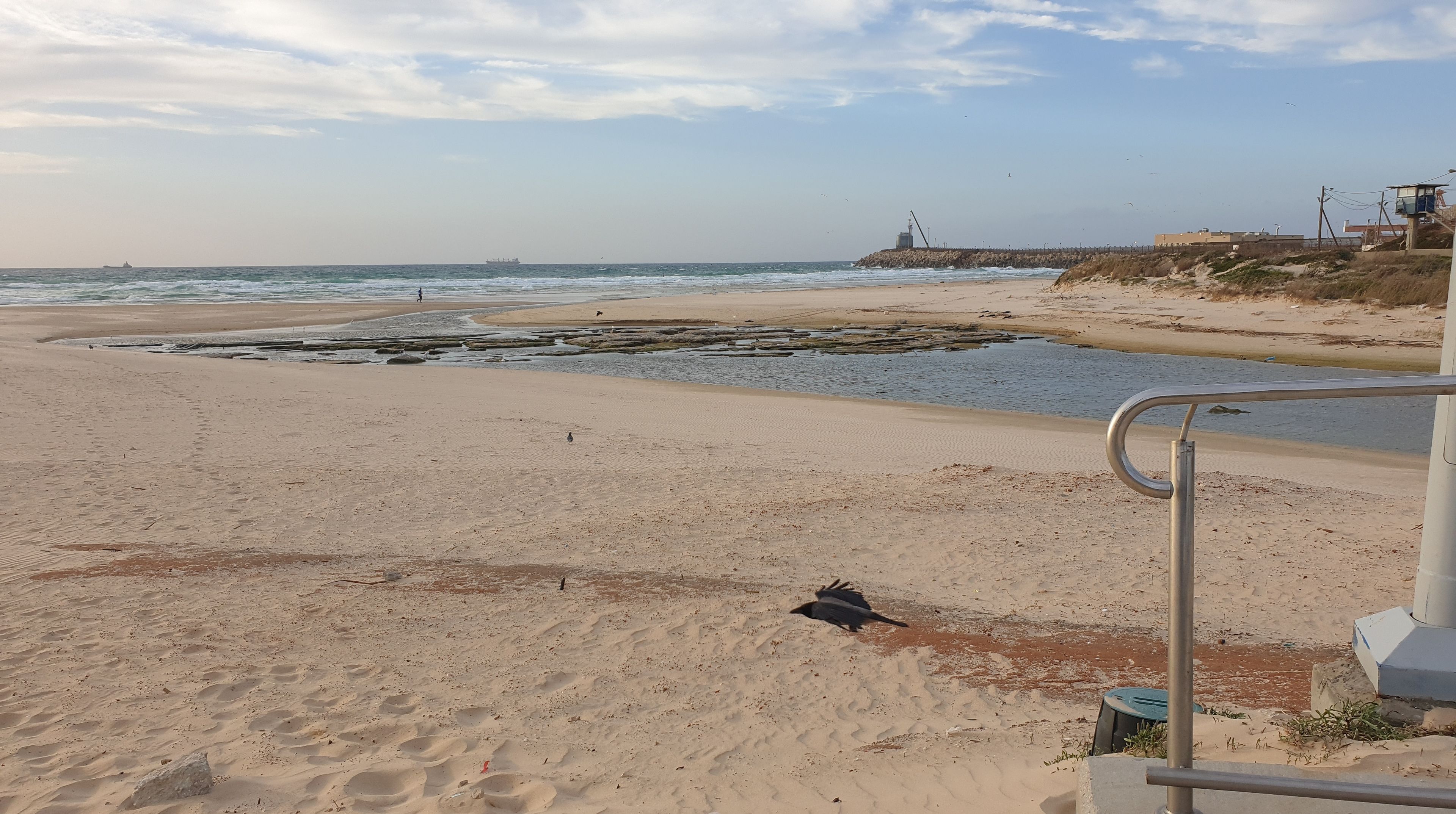 Una playa con un promontorio rocoso, un muelle y un faro a lo lejos.