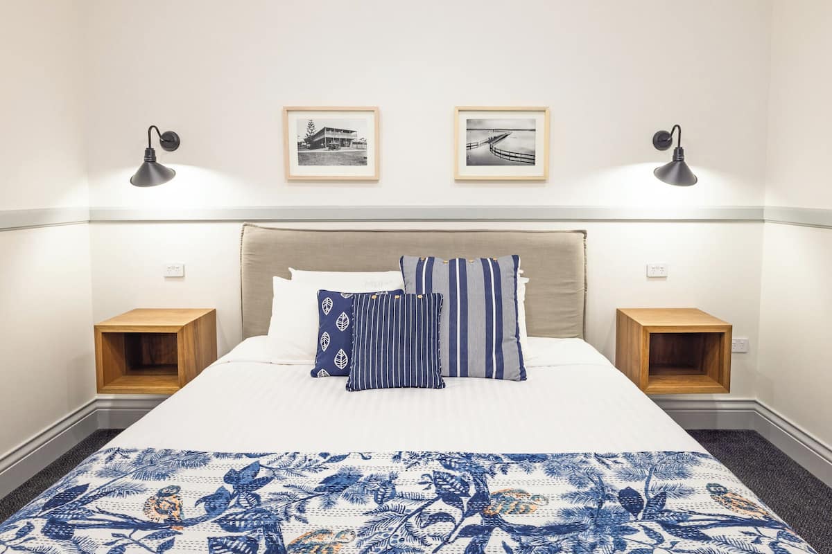 Una cama bien hecha con una colcha estampada, dos mesitas de noche, un cabecero y dos cuadros enmarcados en la pared.