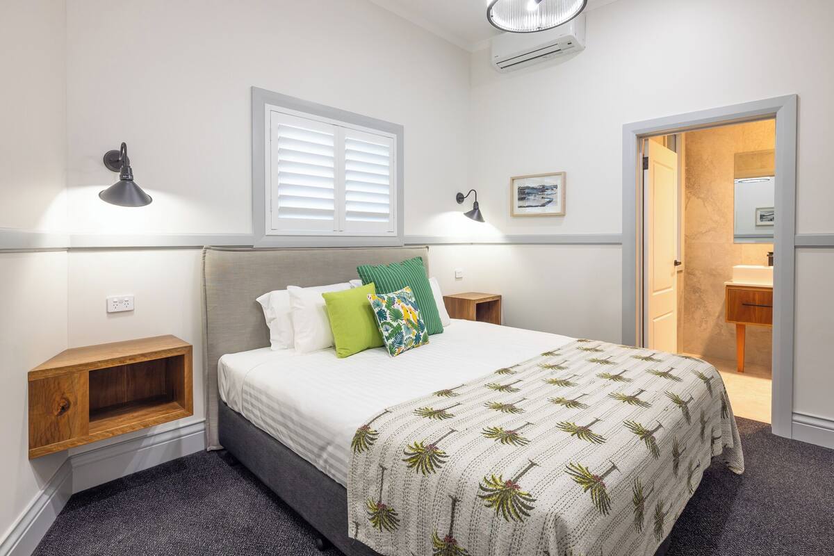 Un dormitorio con cama, lámparas de noche, una mesita de noche de madera, aire acondicionado de pared, una ventana con persianas y una puerta que da al baño.