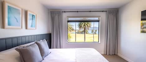 A hotel room with a large bed, a ceiling fan, a window with a view of palm trees and mountains, and two framed pictures on the wall.
