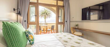 Ein Schlafzimmer mit Bett, grün gestreiften Kissen, einem Fenster mit Vorhängen, einem Balkon mit Tisch und Stühlen sowie einem Wand-Fernseher.