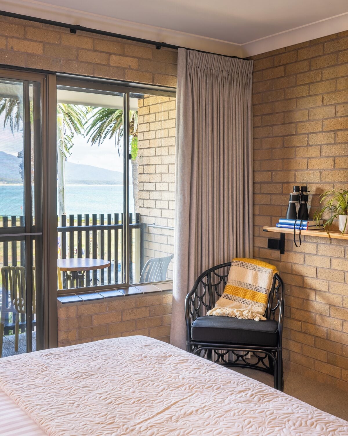 A hotel room with a large bed, a chair with a patterned cushion, a shelf with binoculars, and a view of the sea through a glass door.