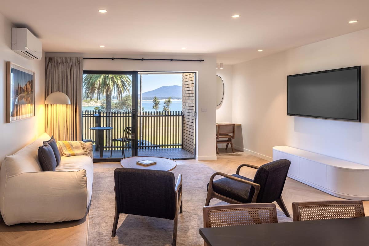 Una sala de estar moderna con un sofá, sillas, una mesa de centro, un televisor y un balcón con vistas al mar y a las montañas.