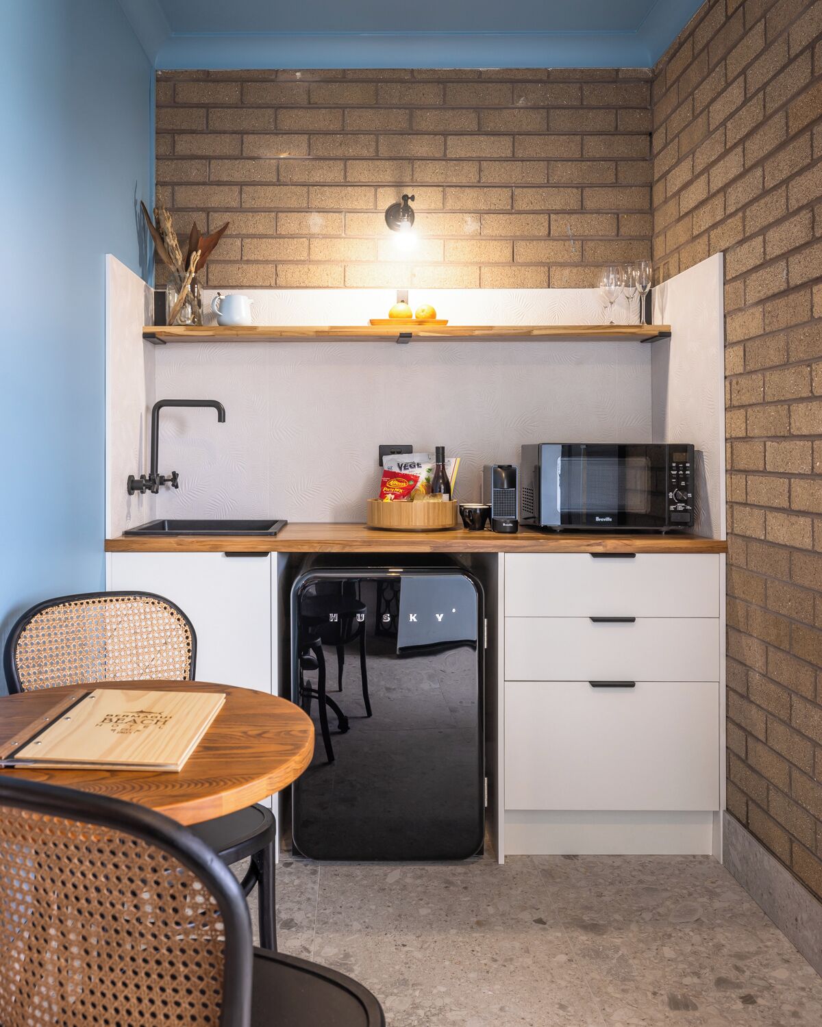 A compact kitchen with a sink, microwave, and a small refrigerator. There is a wooden table with a folder and a chair.