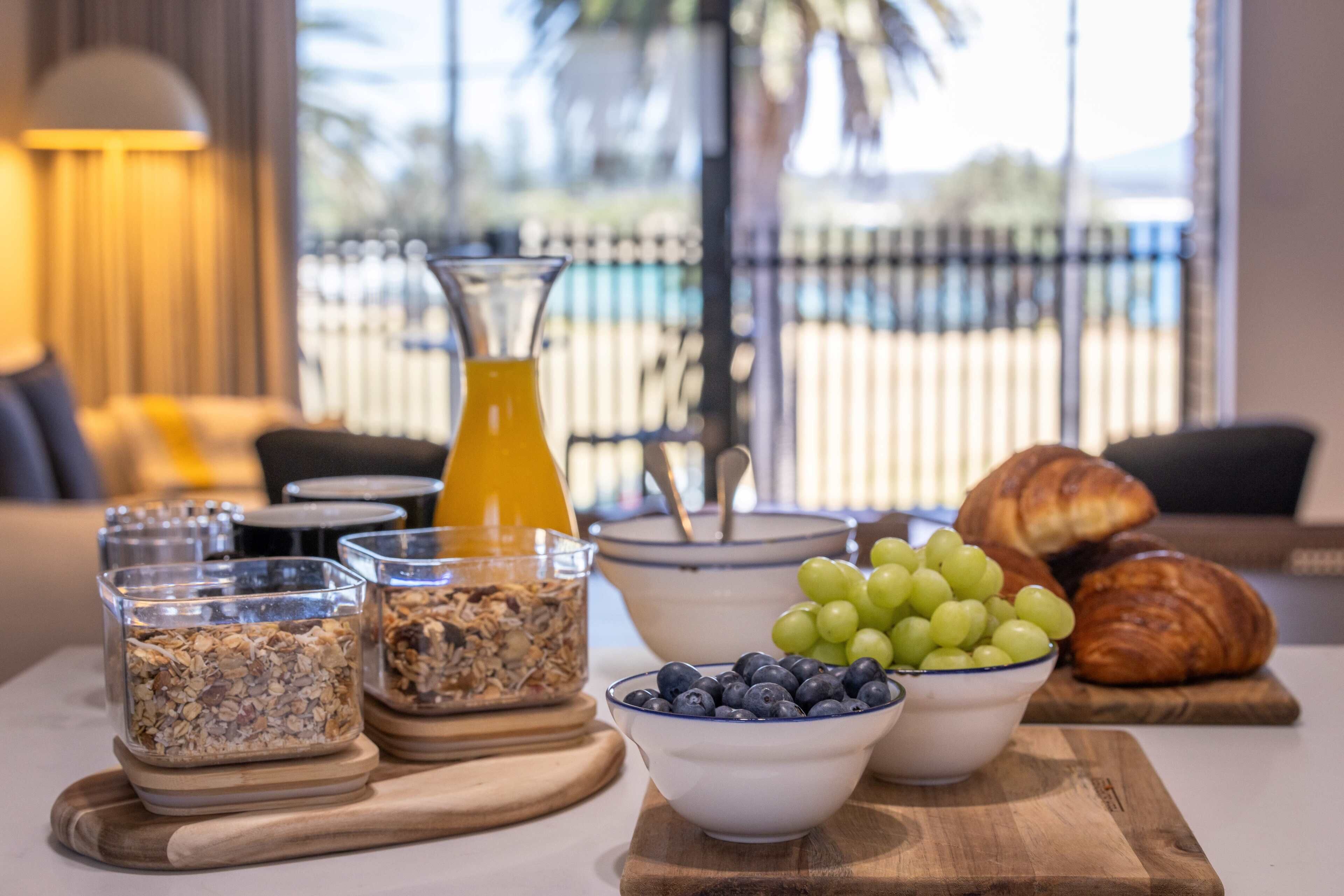 Une table de petit-déjeuner avec des bols de granola, des myrtilles et des raisins, un verre de jus d’orange et des croissants.
