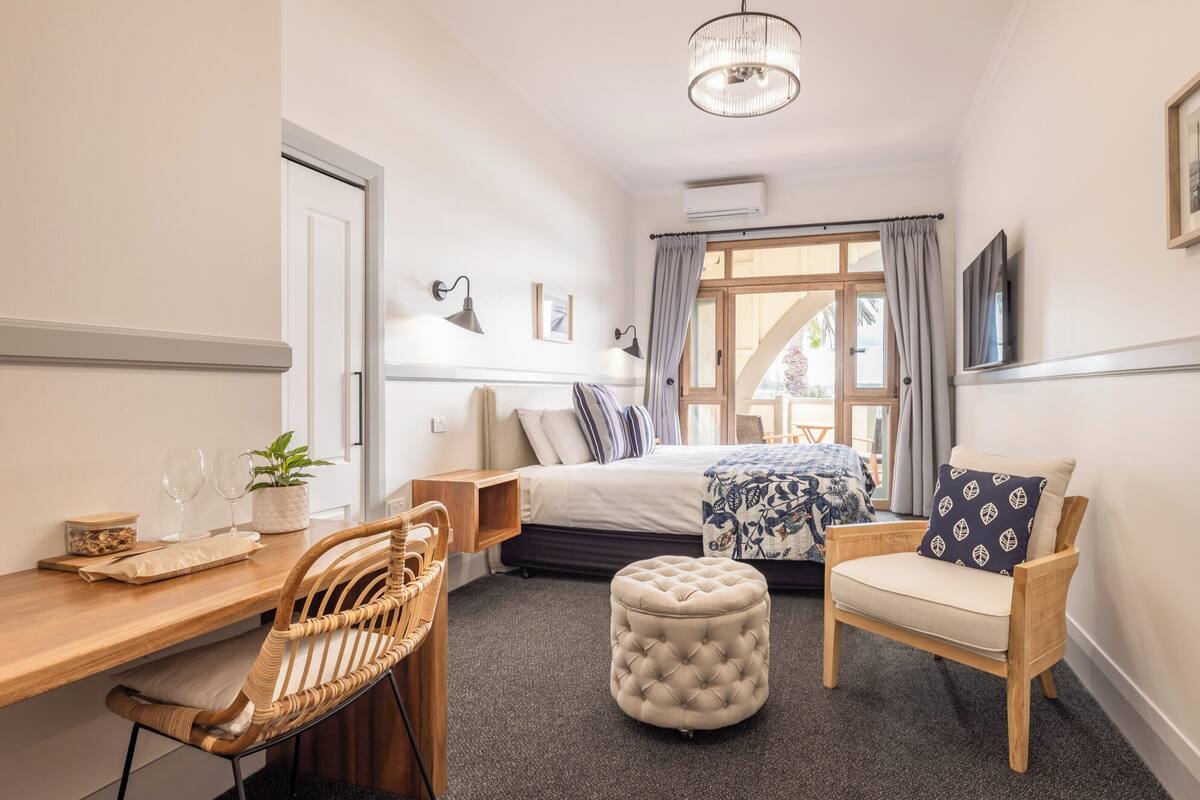 Habitación de hotel pequeña con cama, escritorio, silla y un pequeño reposapiés.