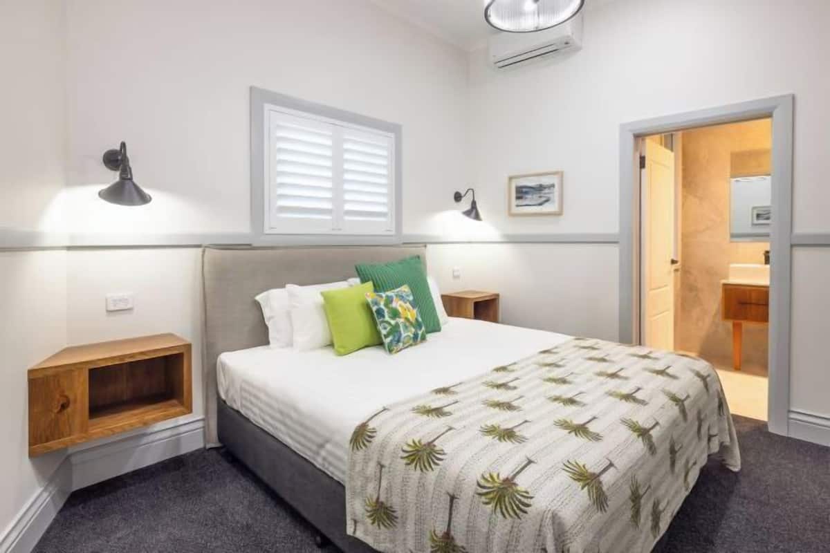 Un dormitorio con cama, mesitas de noche, una ventana con persianas, aire acondicionado de pared y una puerta que da a otra habitación.
