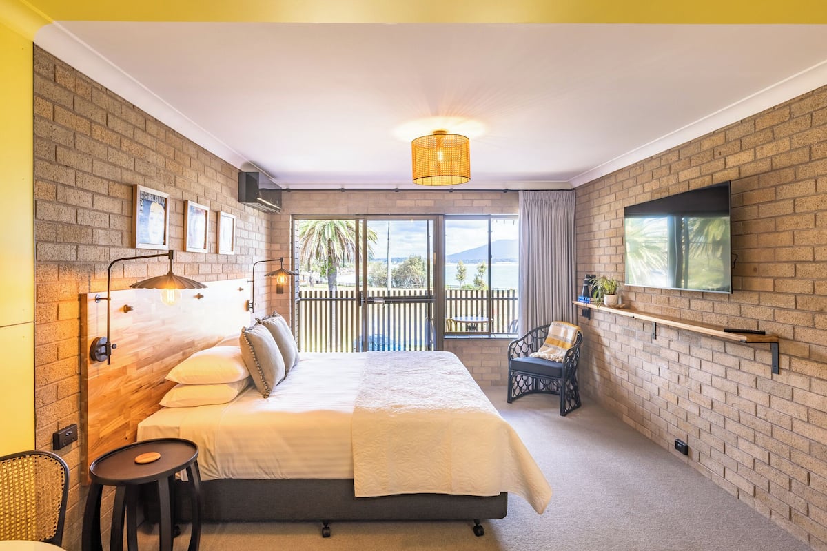 Habitación de hotel con una cama grande, televisión montada en la pared, balcón con vistas y un pequeño rincón de descanso.