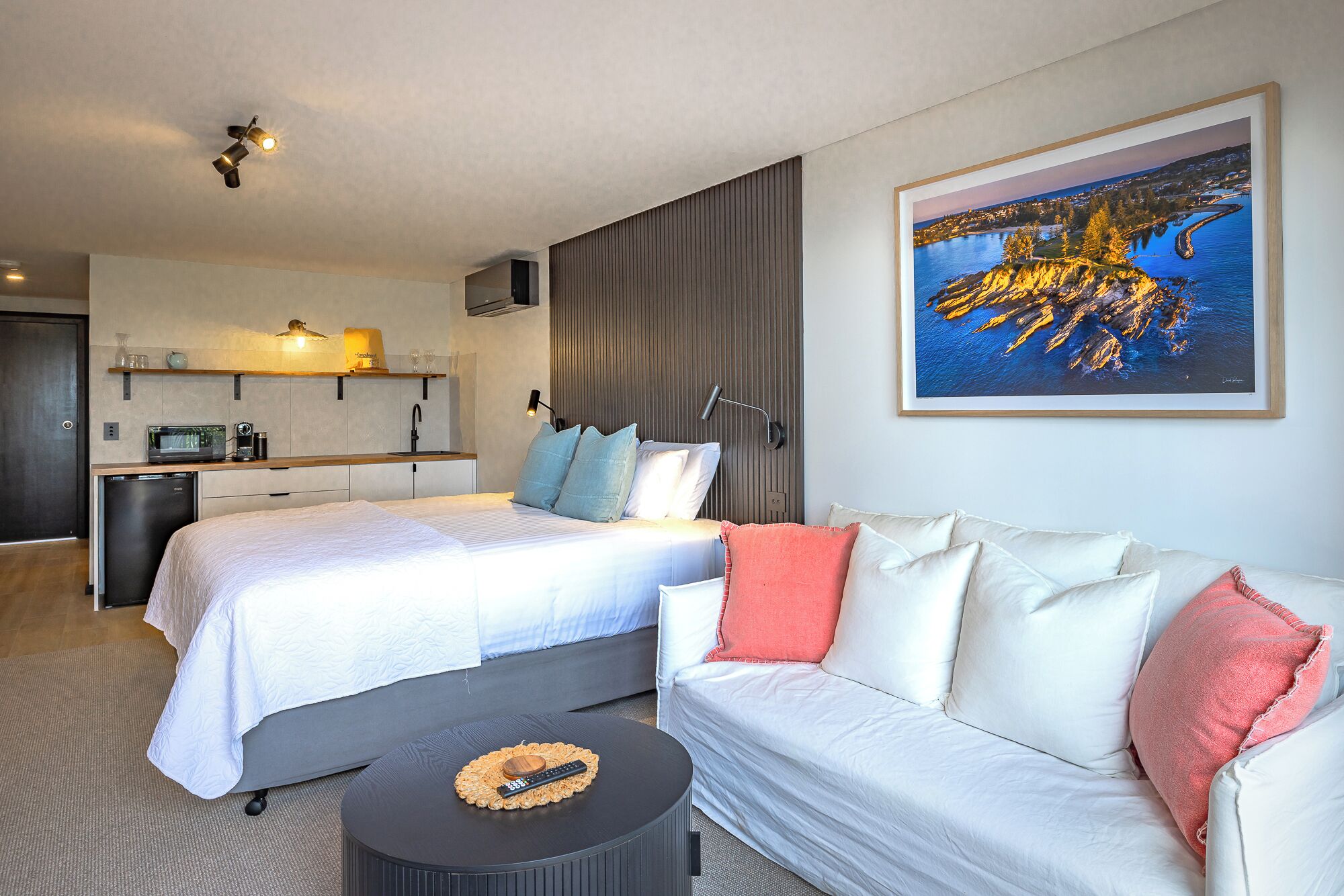 A hotel room with two beds, a sofa, a small table with a remote, a kitchenette, and a framed landscape painting.