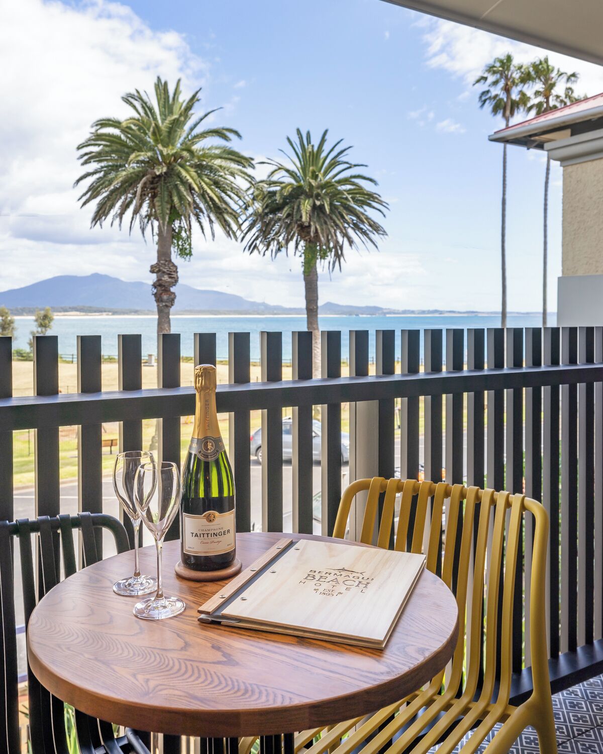 A table with a bottle of champagne and two glasses on a balcony overlooking a beach with palm trees and mountains in the distance.
