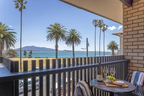 A balcony with a view of the beach and ocean, a table with a bottle of wine, glasses, and grapes, and a chair with a towel. - Bermagui Beach Hotel (Bermagui)