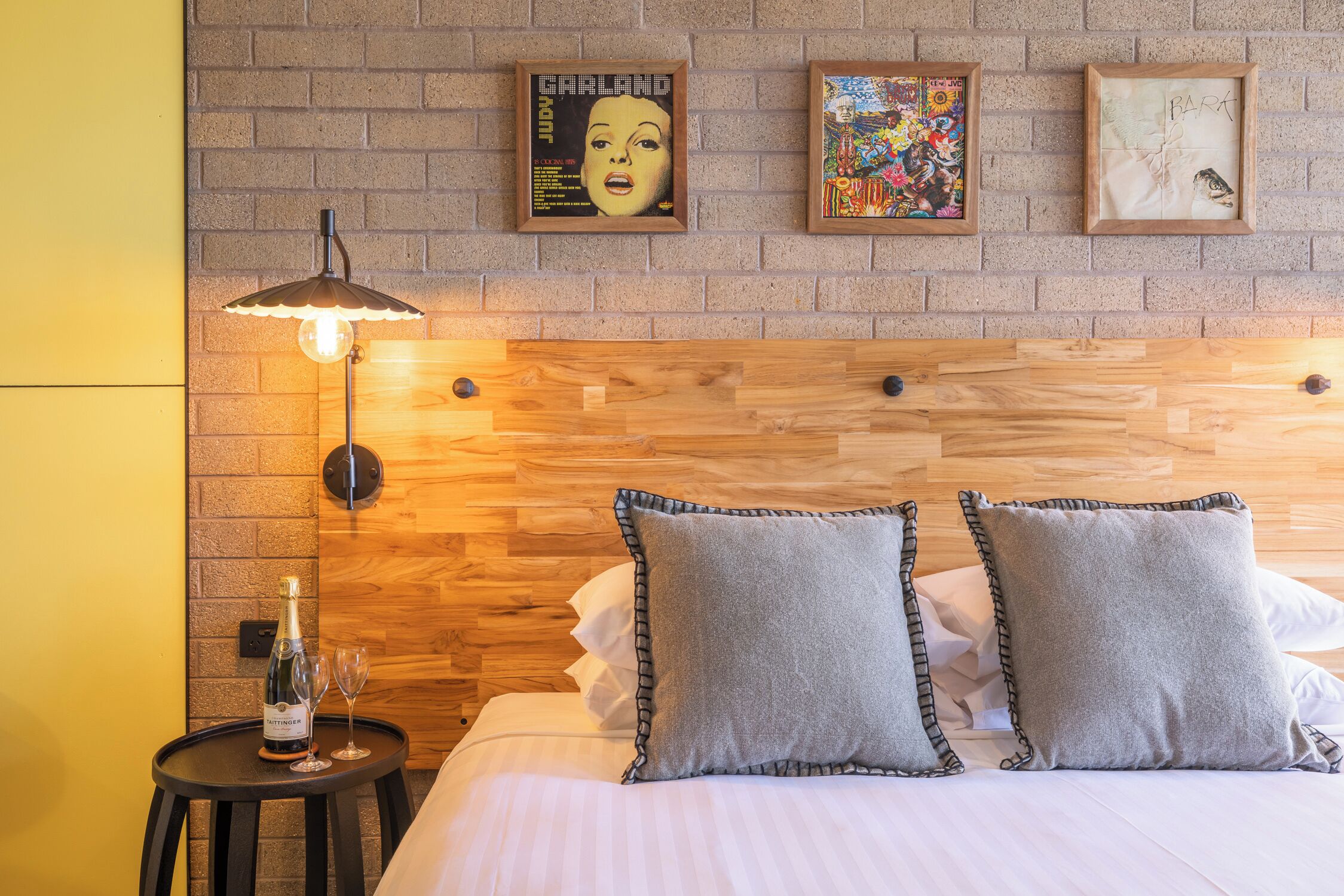 A hotel room with a brick wall, wooden headboard, and a small table with a bottle and glasses.