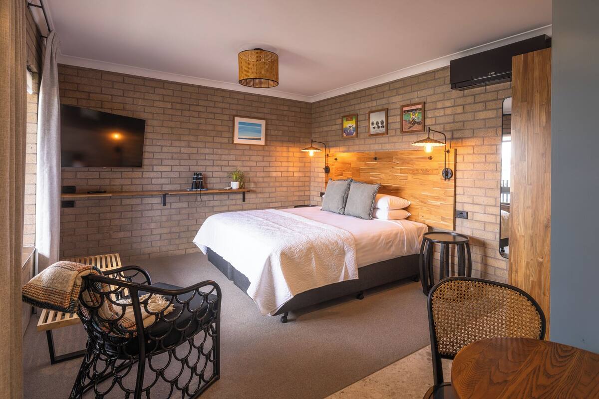 Un dormitorio con pared de ladrillo, una cama con ropa blanca, un televisor montado en la pared, cabecera de madera y una pequeña mesa con sillas.