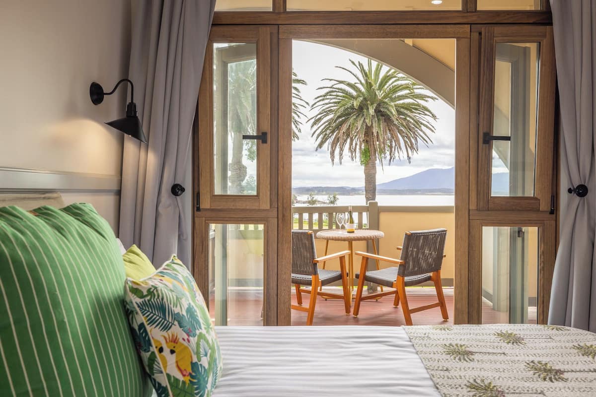 Una habitación con cama, vistas a un balcón con mesa y sillas, y un ventanal con cortinas.