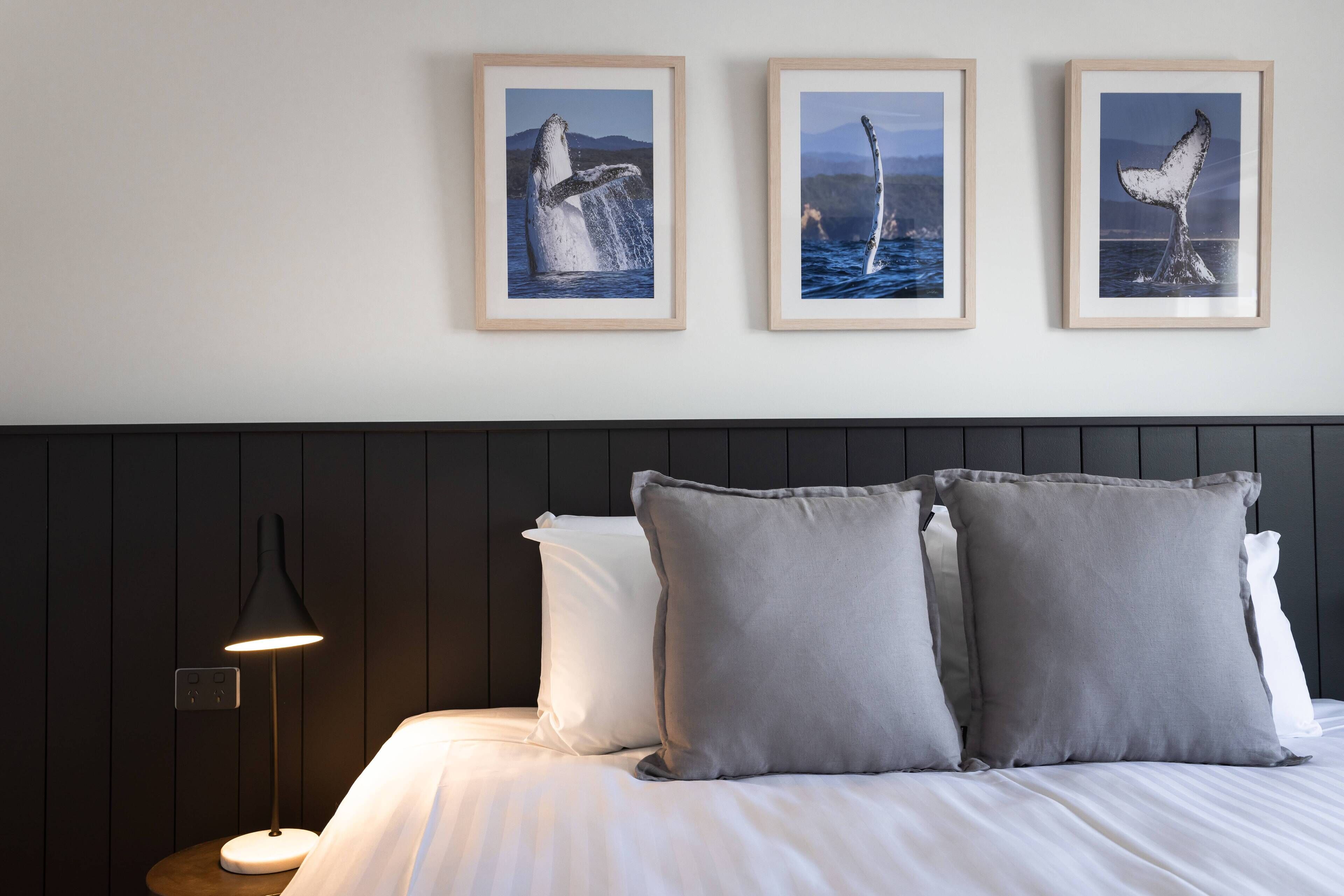 A neatly made bed with white linens, two gray pillows, and a black lamp on a wooden bedside table. Three framed whale tail photographs are hung on the wall above the bed.