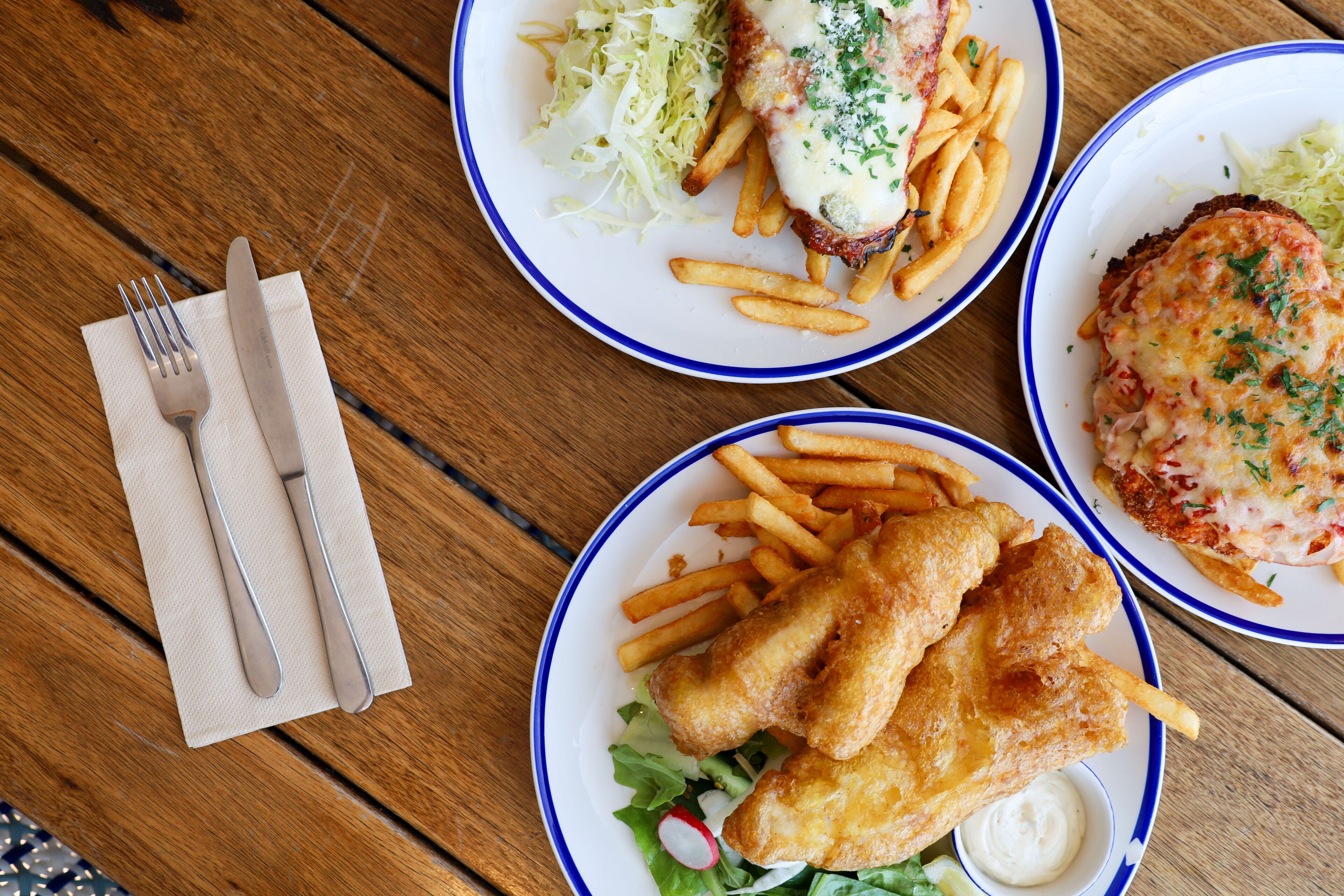 A wooden table with three plates of food, including fish and fries, and a fork and knife set on a napkin.