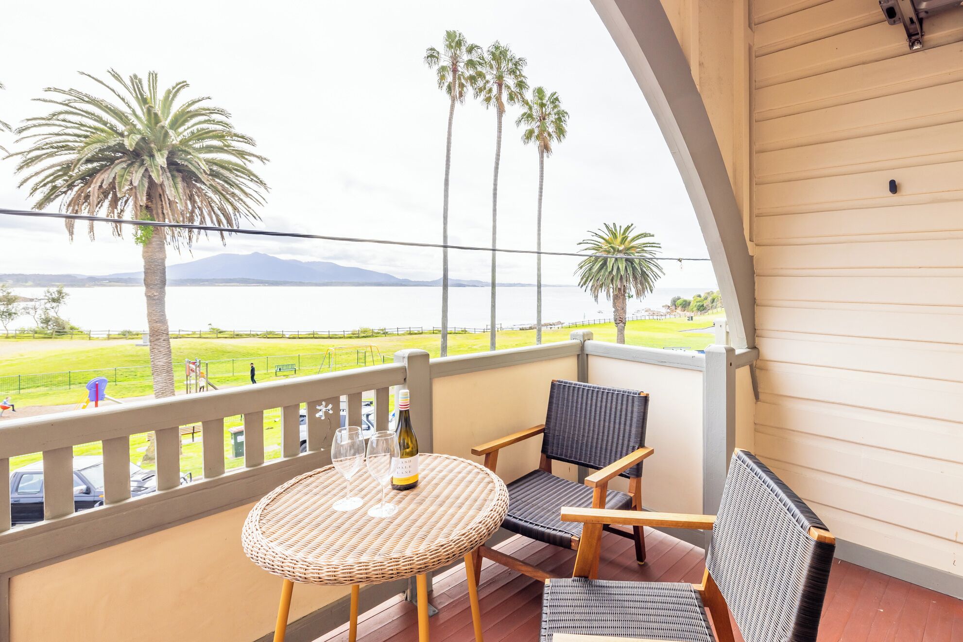 A balcony with wicker furniture, a bottle of wine, and a view of a grassy area with palm trees and a body of water.