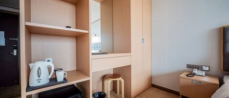 Una habitación de hotel compacta con kitchenette, un taburete pequeño, un cesto de basura, una mesita de noche con teléfono y televisor.