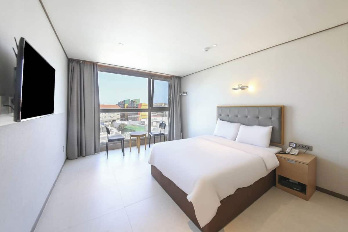 Hotelkamer met een groot bed, een televisie, een bureau met telefoon en uitzicht op gebouwen door een groot raam.