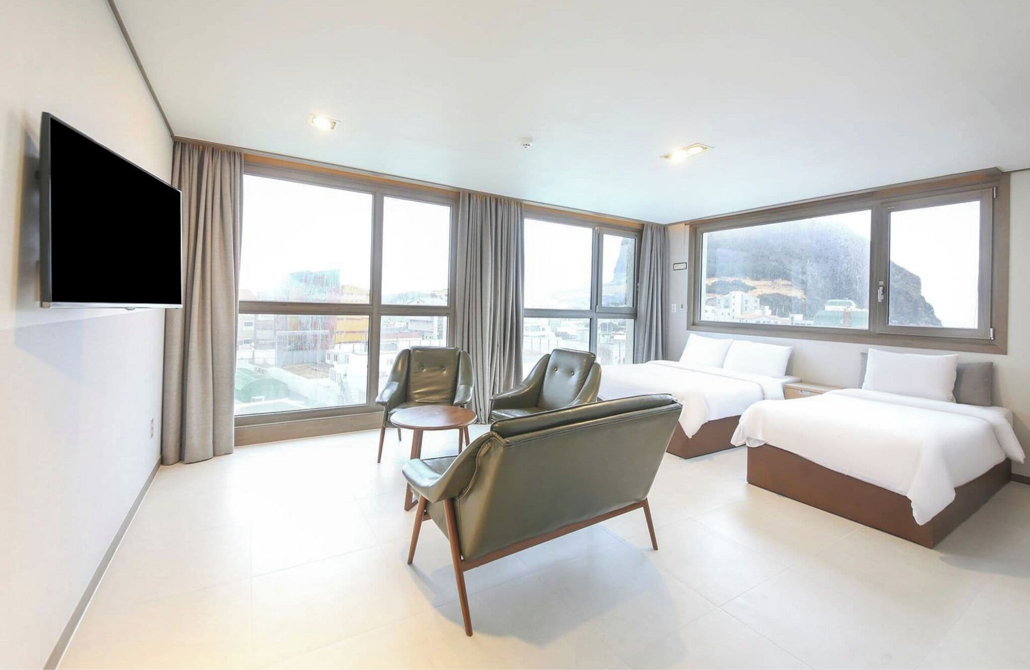 A hotel room with a bed, two chairs, a TV, and large windows offering a city view.