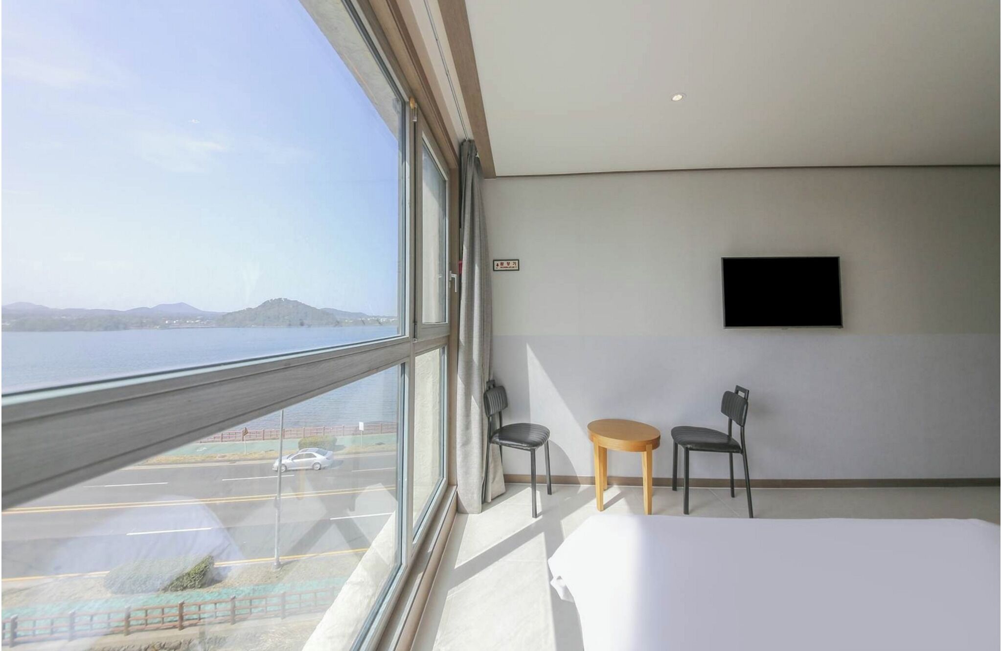 Een moderne hotelkamer met een groot raam, met uitzicht op een waterlichaam en heuvels in de verte. Er is een bed, een klein tafeltje, twee stoelen en een flatscreen televisie bevestigd aan de muur.