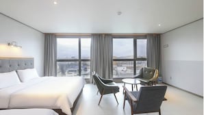 Habitación de hotel con cama, dos sillas, una mesita y vistas a la ciudad a través de amplias ventanas.