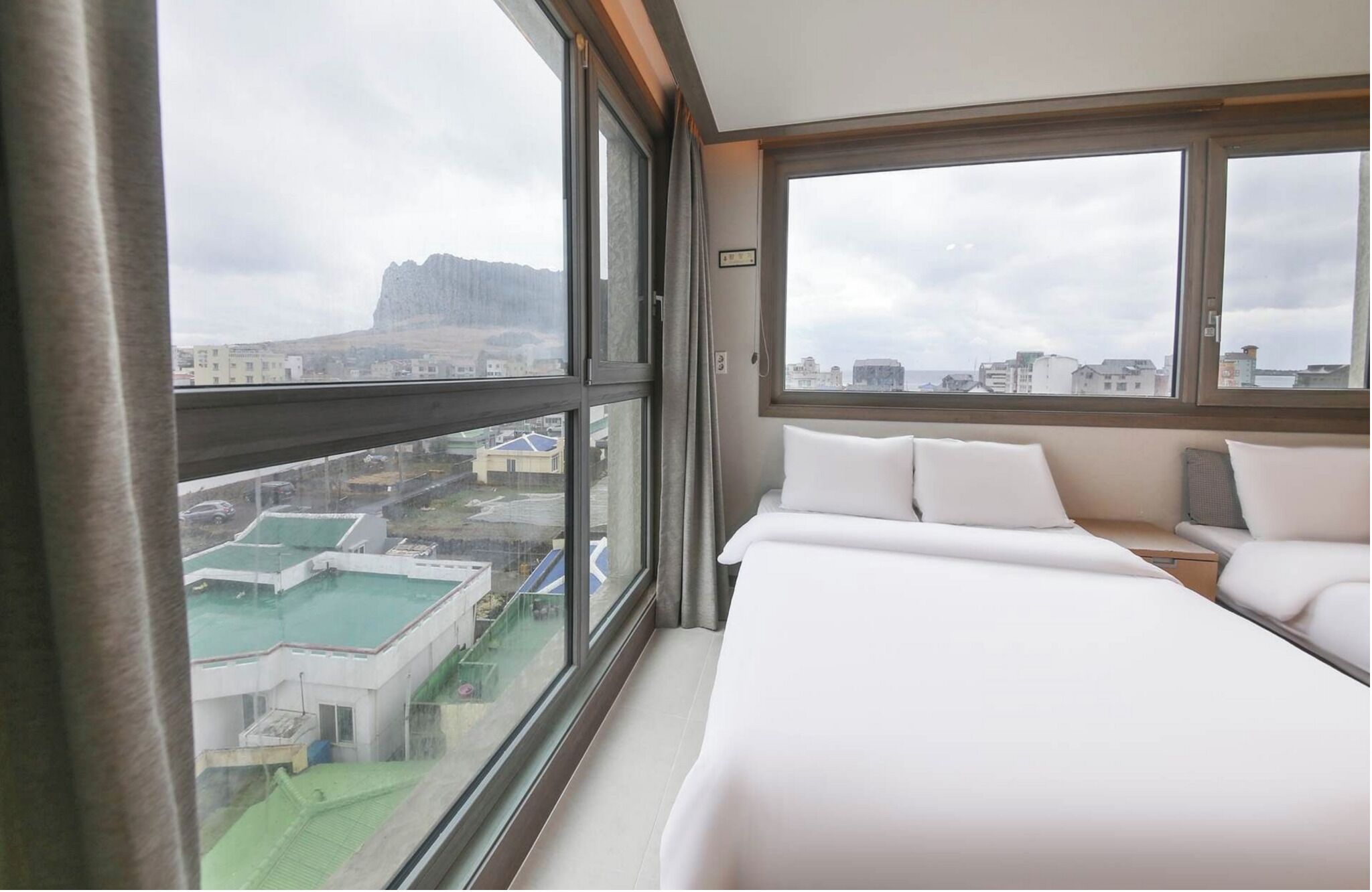 A hotel room with a large window, a bed, a bedside table, and a view of buildings and a mountain.