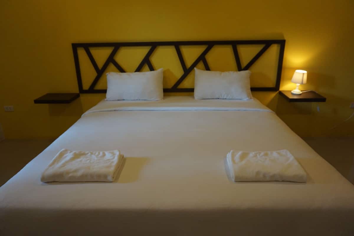 A neatly made bed with pillows and folded towels, a bedside table with a lamp, and a wall with a geometric pattern.