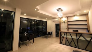 A modern hotel lobby with a reception desk, a dining area with a table and chairs, and a large glass wall offering a view of the city. - Somsamai Style Apartment (Khon Kaen)