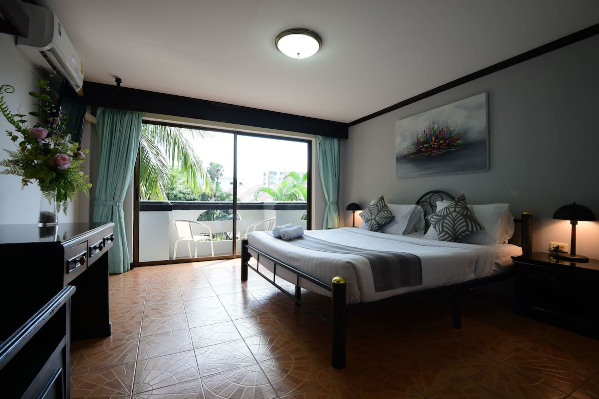 A bedroom with a bed, a nightstand, a lamp, a window with curtains, a balcony, and a view of greenery outside.