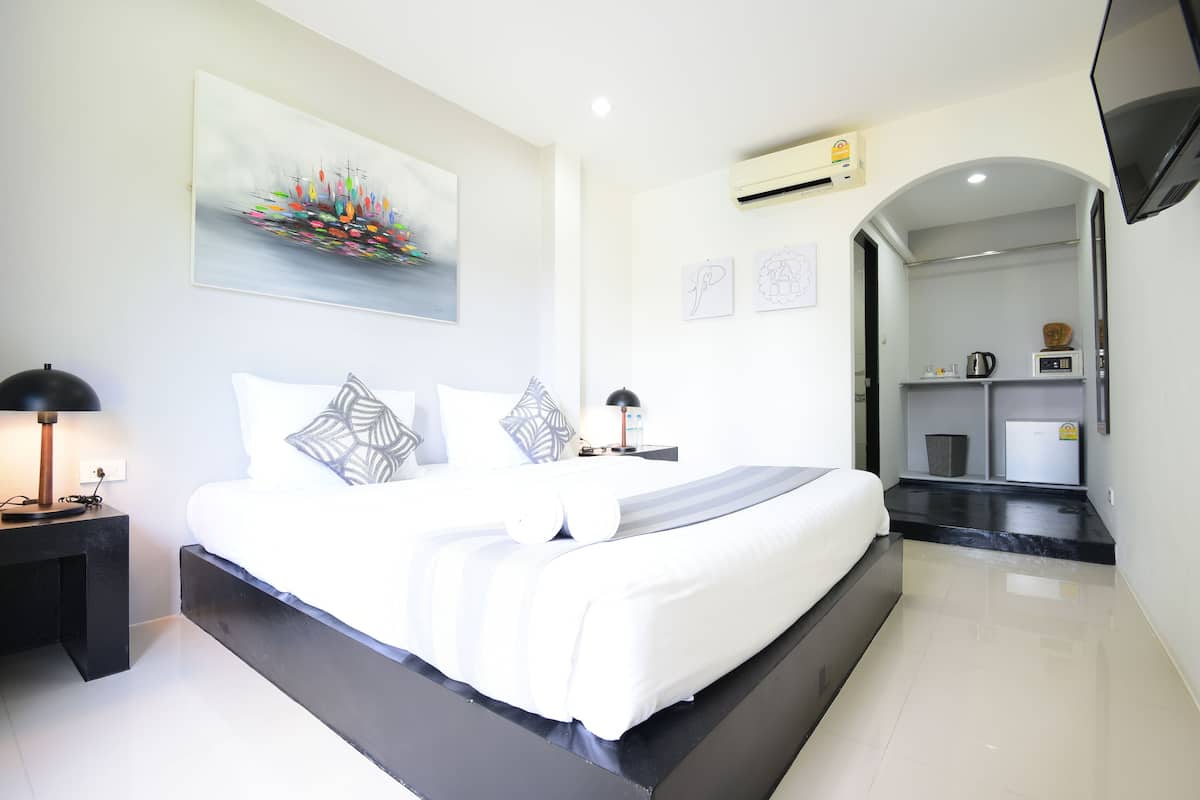 A modern hotel room with a large bed, a bedside table with a lamp, a flat-screen TV, and a small kitchenette area.
