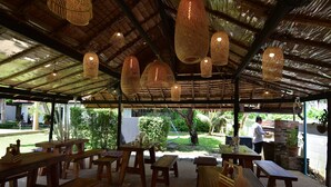 Un patio al aire libre con mesas y bancos de madera, lámparas colgantes de tejido artesanal y una estructura de techo de paja.