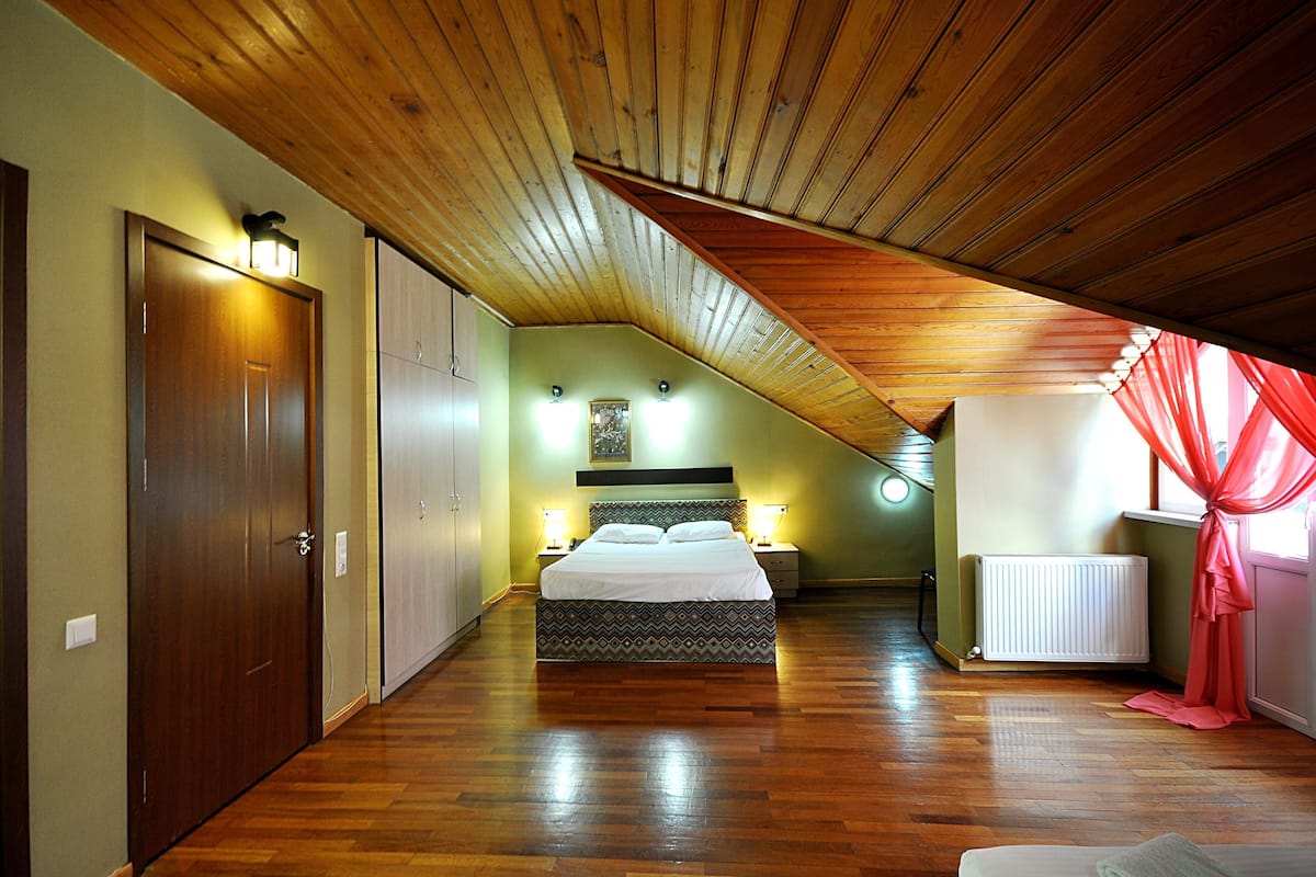 Un dormitorio con techo de madera, cama con cabecera estampada, lámparas de noche y ventana con cortinas rojas.