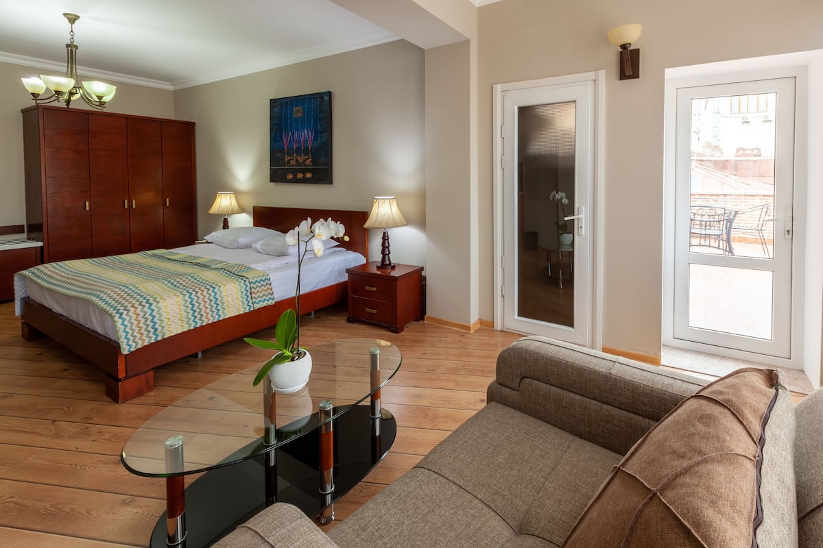 A bedroom with a bed, bedside tables, a nightstand, a sofa, a glass coffee table, a potted plant, a painting, and a sliding glass door leading to a balcony.