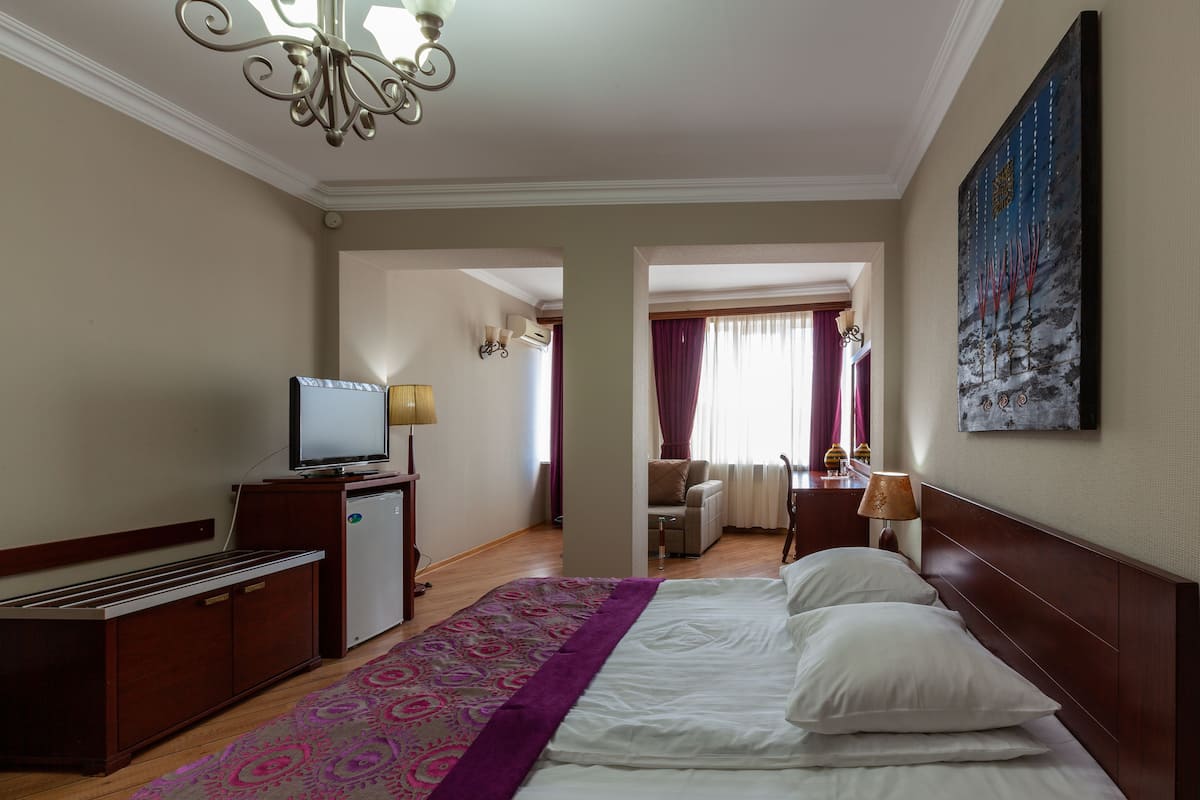 A hotel room with a bed, a desk with a television, a small refrigerator, a lamp, and a painting on the wall.