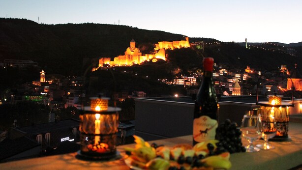 A table with fruit and a bottle of wine, overlooking a lit-up historic town with a prominent castle-like structure on a hill. - Hotel Penthouse (Tbilisi)