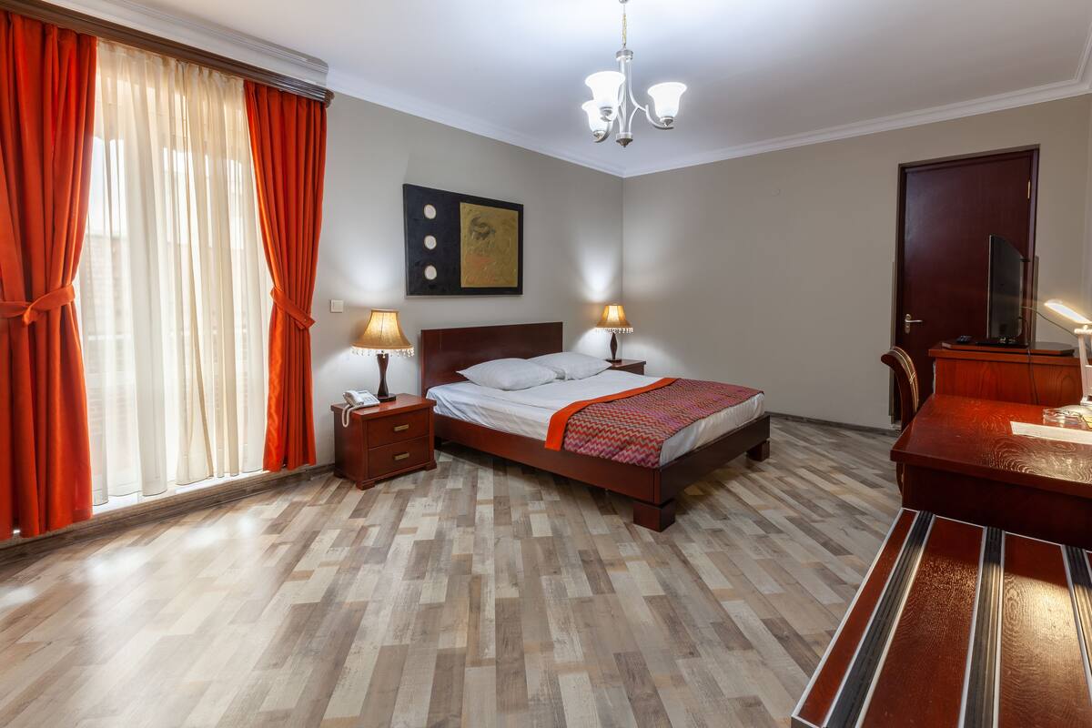 A hotel room with a wooden bed, two bedside tables with lamps, a large window with curtains, a painting on the wall, and a door with a mirror and a small table.