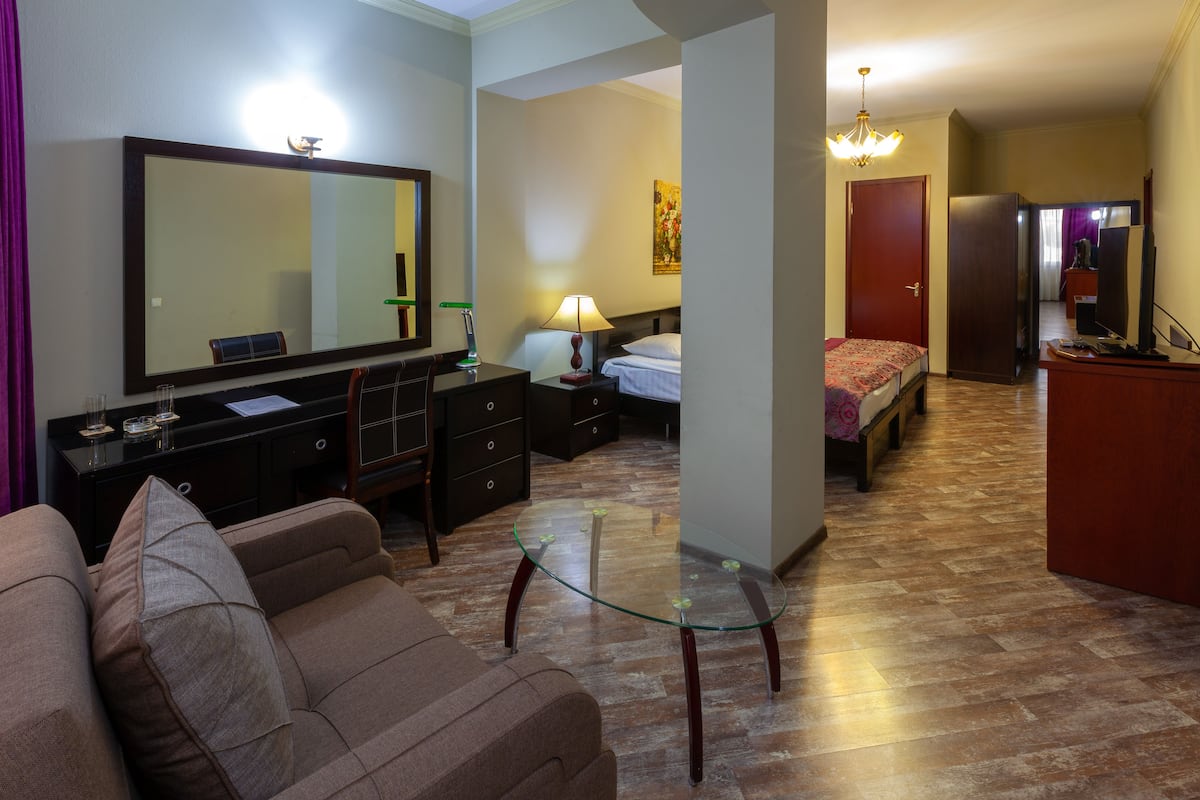 A hotel room with a large mirror, a bed, a desk with a lamp, a sofa, and a glass coffee table.