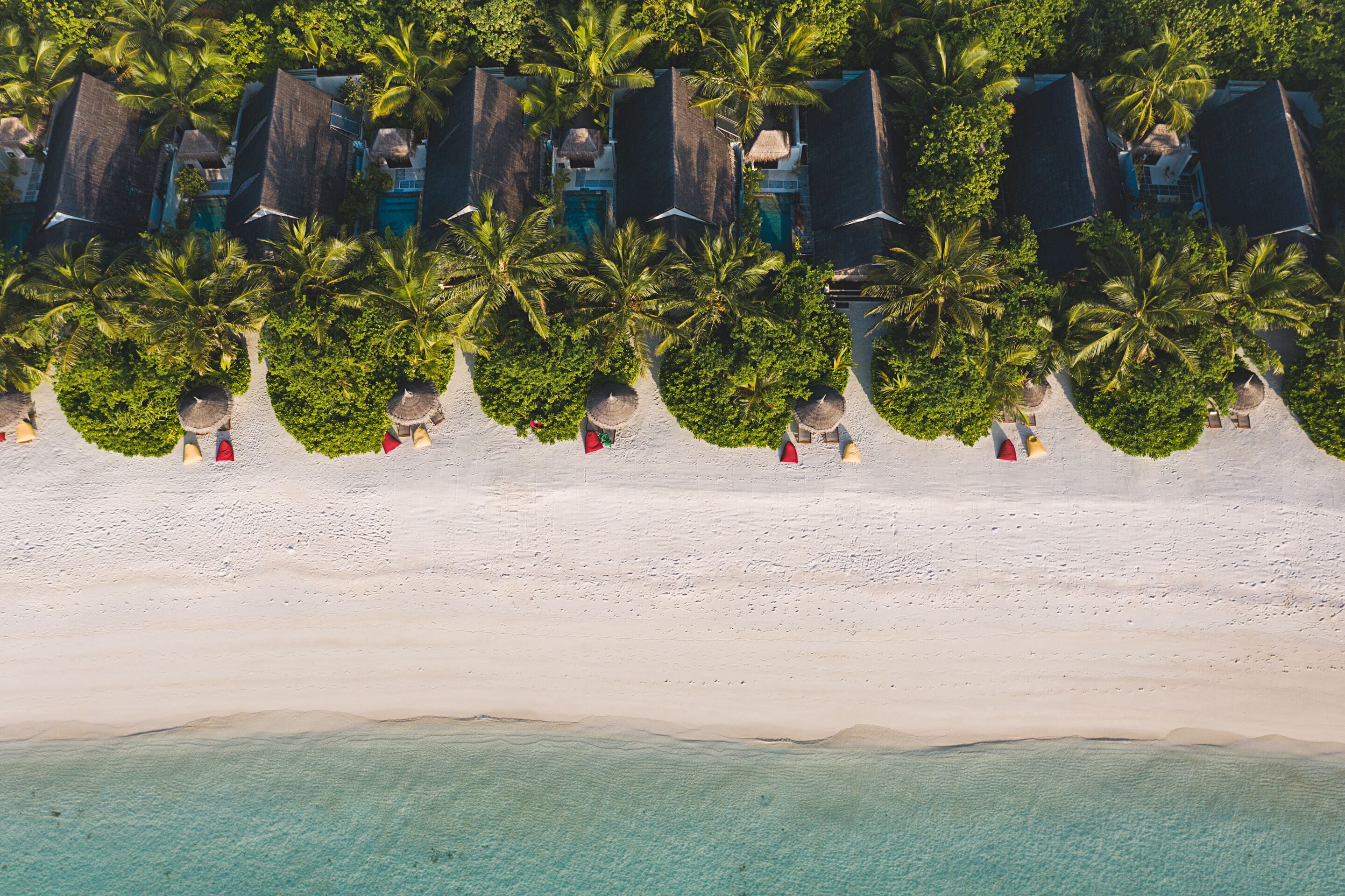Vista aérea de un complejo turístico en la playa con filas de bungalows sobre el agua, palmeras y una playa de arena.