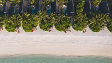 Vista aérea de un complejo turístico en la playa con filas de bungalows sobre el agua, palmeras y una playa de arena.